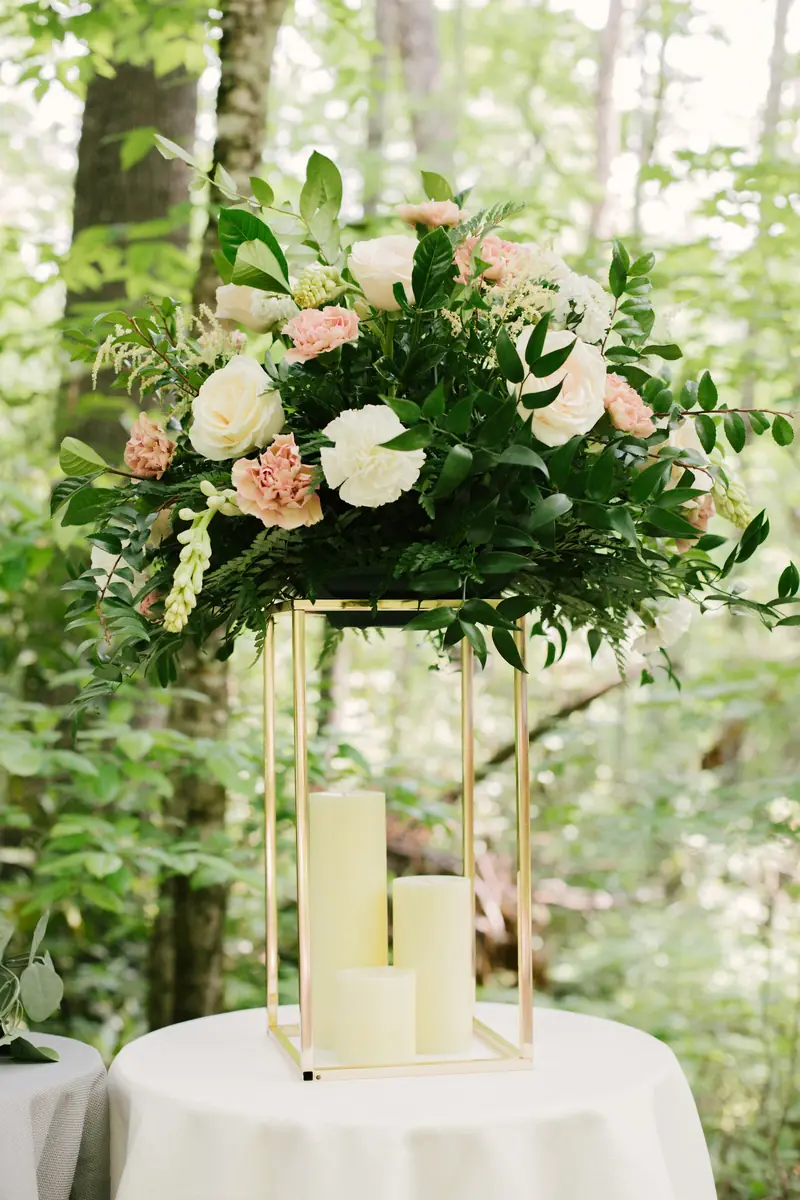 A tall gold floral stand with blush roses and greenery above pillar candles