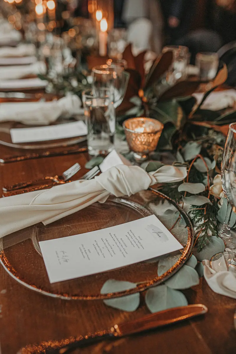 A place setting with a copper-rimmed charger, knotted ivory napkin, eucalyptus and a gold votive